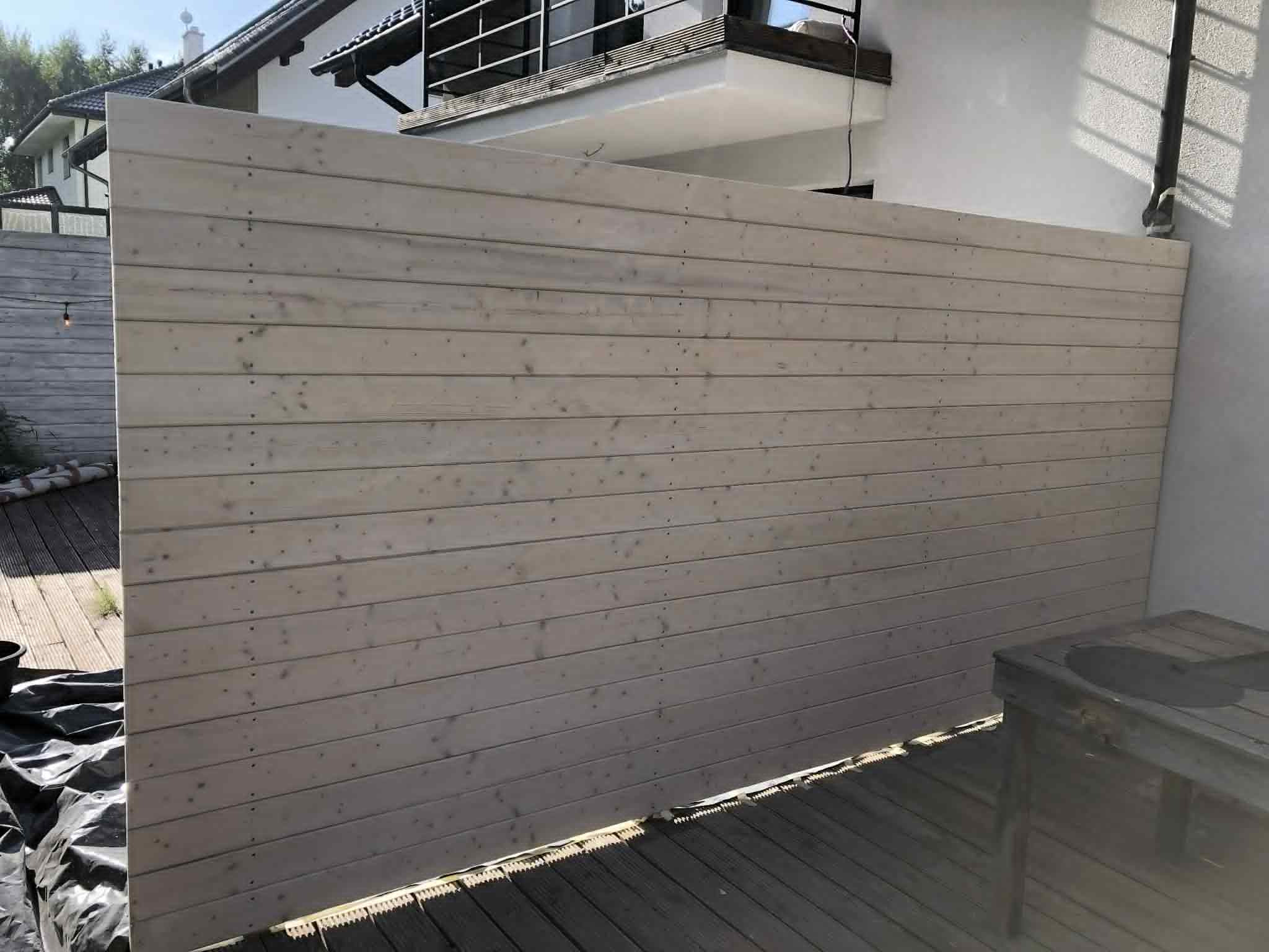 Jasna, drewniana osłona tarasu z desek, widoczna z boku. Częściowo widoczny taras i fragment budynku w tle. Minimalistyczny design.