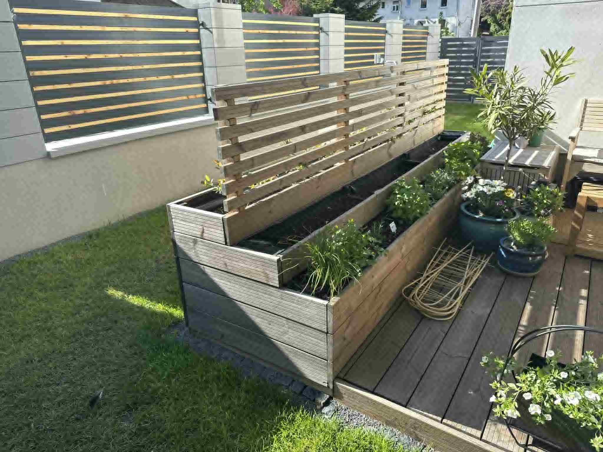 Drewniana, wielopoziomowa rabata warzywna z osłoną, na tarasie z desek. W tle nowoczesne ogrodzenie z poziomych desek. Słoneczny dzień, zadbany ogród.