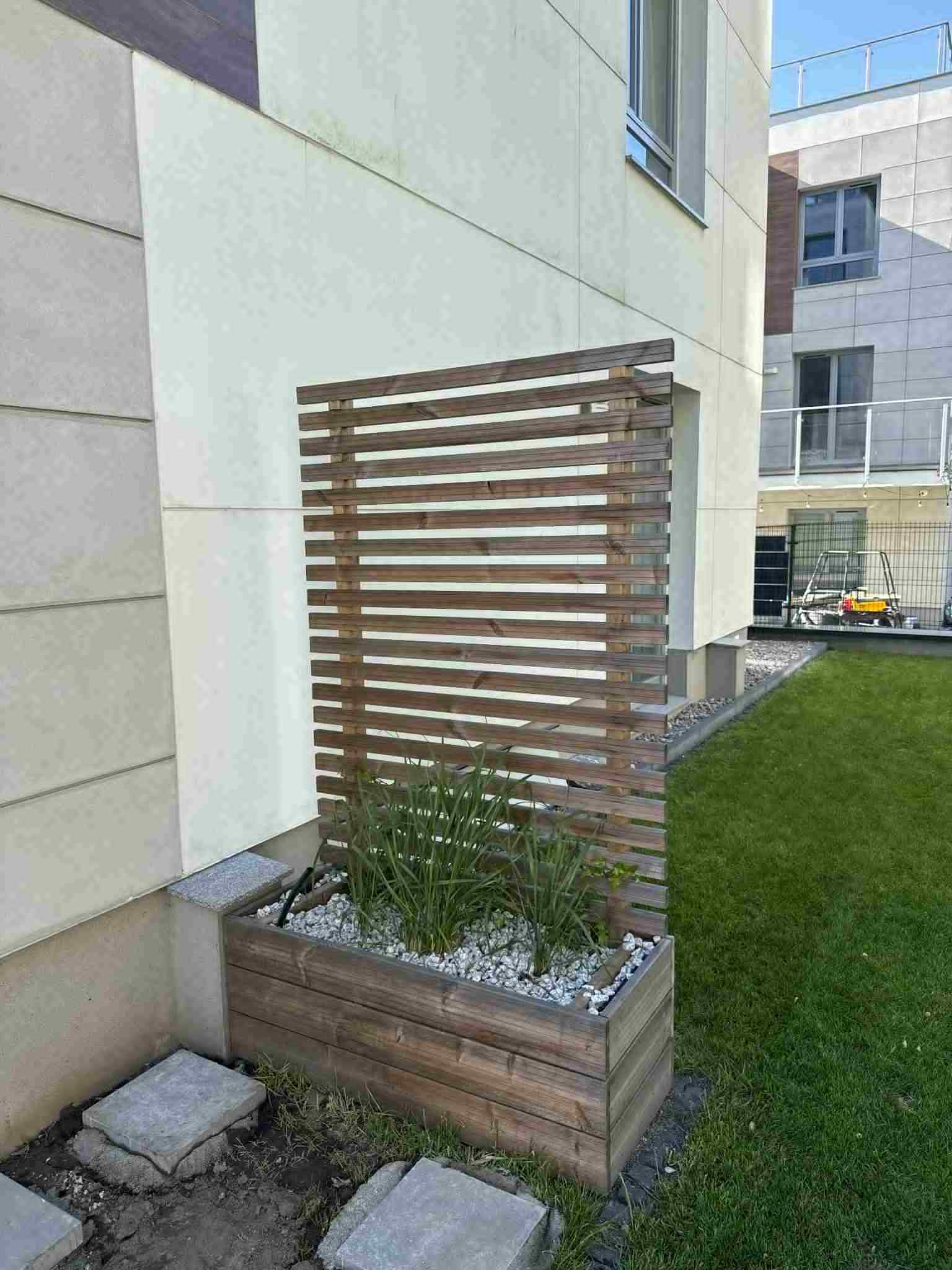 Nowoczesna drewniana ścianka działowa z poziomych listew, połączona z podwyższoną rabatą, wypełniona roślinami i białym żwirem przy budynku.