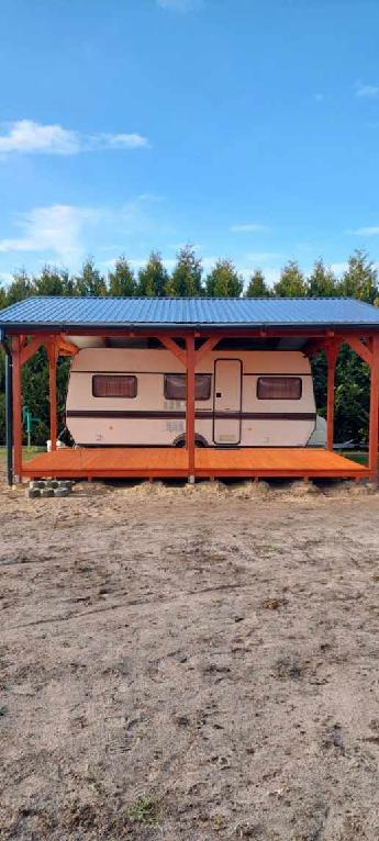 Zadaszona drewniana konstrukcja z niebieskim dachem, pod którą stoi przyczepa kempingowa. Widok na tle zieleni i błękitnego nieba.