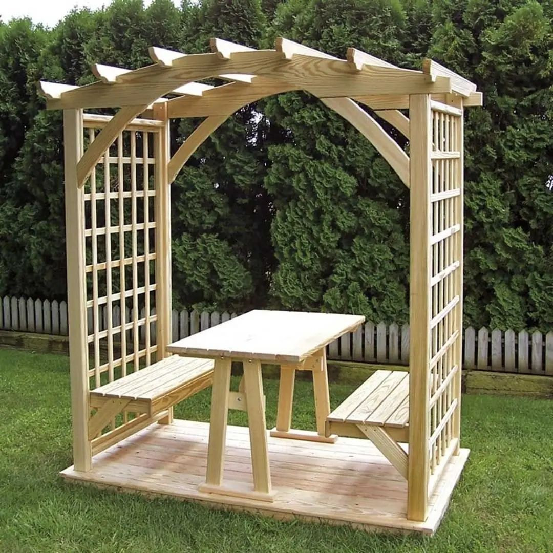 Drewniana altana z ławkami i stołem, zadaszona konstrukcją z łukiem, na drewnianym podeście, w ogrodzie z zielonymi krzewami i białym płotem w tle.