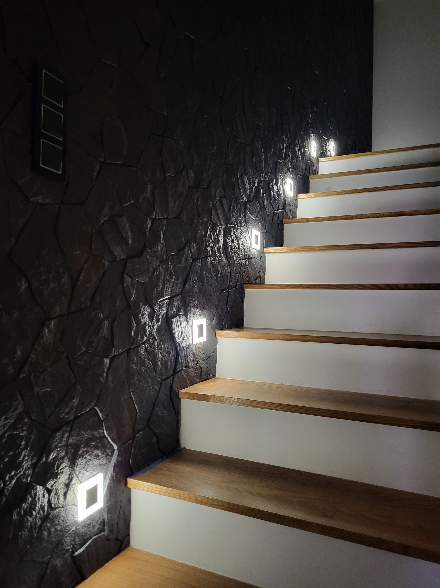Schody z drewnianymi stopniami i oświetleniem LED wbudowanym w ciemną, kamienną ścianę. Nowoczesne wykończenie wnętrza.