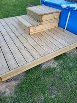 Drewniany taras z dwustopniowymi schodkami przy basenie ogrodowym, widok z góry na jasne deski i zieloną trawę w tle. Świeżo zaimpregnowane drewno.