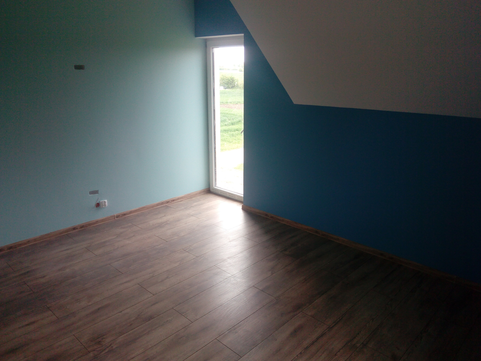 Pokój po remoncie: ciemne panele, ściany pomalowane na dwa kolory (błękit i granat), białe okno z widokiem na zieleń. Widoczne gniazdka elektryczne i fragment poddasza.