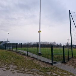 Ogrodex - Otwarta furtka w zielonym ogrodzeniu panelowym otaczającym boisko sportowe, widok na trawę, bramkę i oświetlenie boiska w pochmurny dzień.