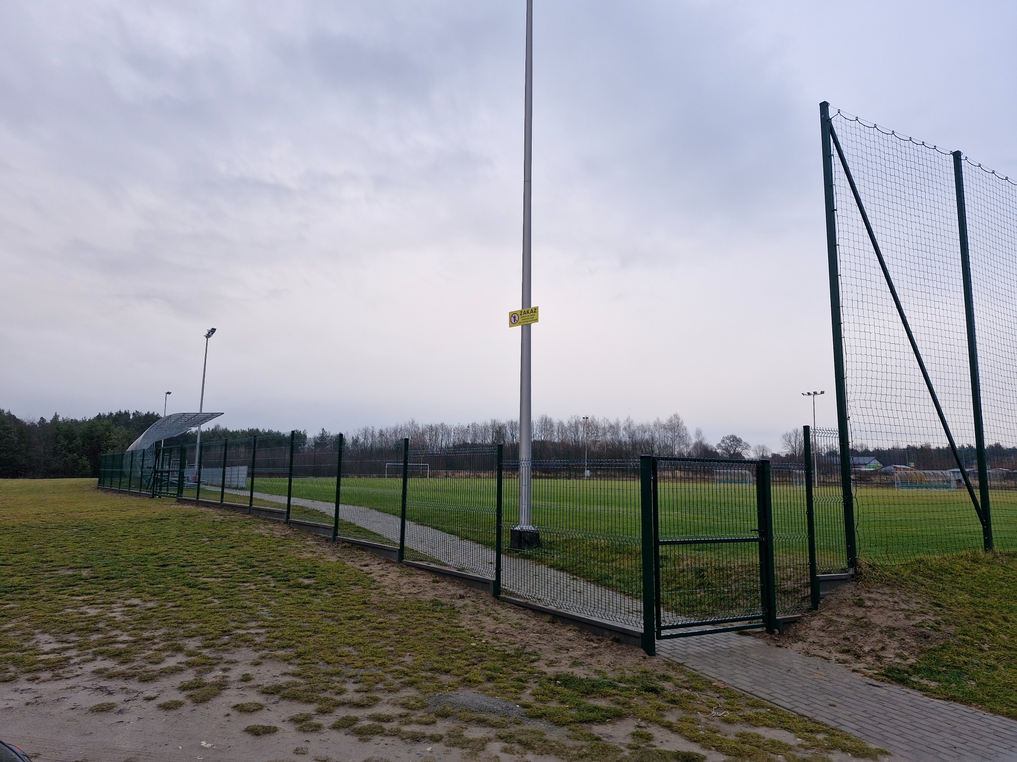Otwarta furtka w zielonym ogrodzeniu panelowym otaczającym boisko sportowe, widok na trawę, bramkę i oświetlenie boiska w pochmurny dzień.