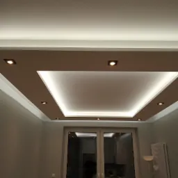 Podwieszany sufit z oświetleniem LED punktowym i taśmowym, widok z dołu na nowoczesny design w jasnym salonie z widocznym fragmentem okna balkonowego.