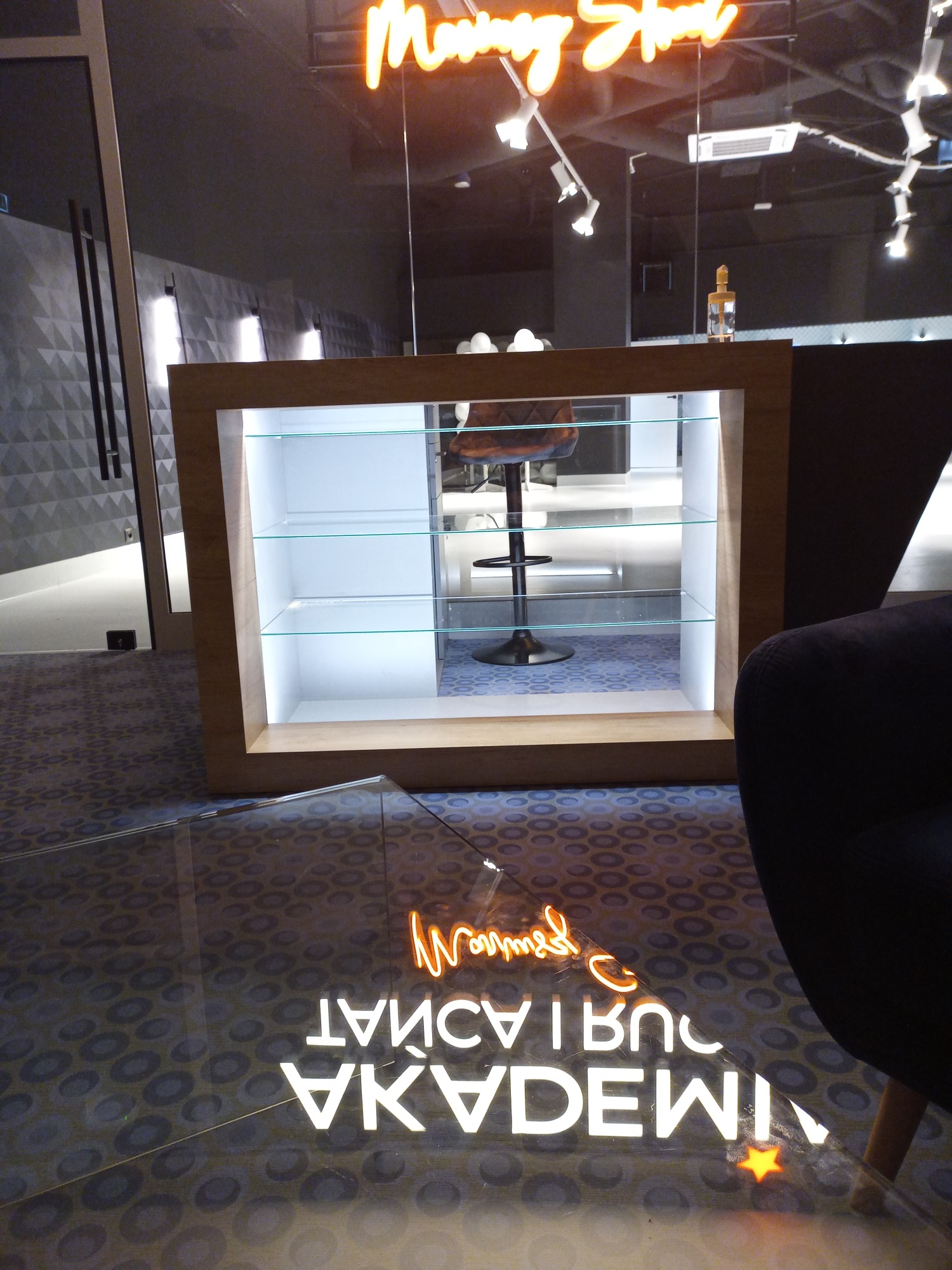 Nowoczesne wnętrze biurowe z podświetlanym neonem na szklanej przegrodzie, widok na recepcję z drewnianą obudową i szklanymi półkami, odbicie neonu na podłodze, ciemna tapicerka sofy w kadrze.