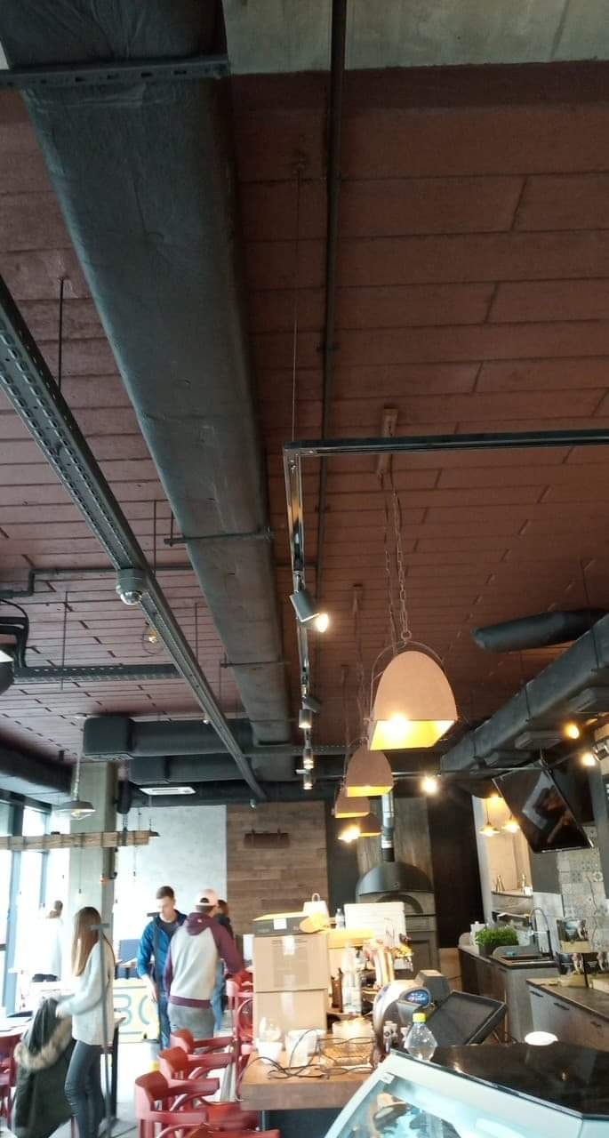 Wnętrze kawiarni z widocznymi elementami instalacji elektrycznej: rurami, przewodami i punktowymi reflektorami na szynoprzewodach, zawieszonymi lampami oraz kamerą monitoringu pod sufitem.