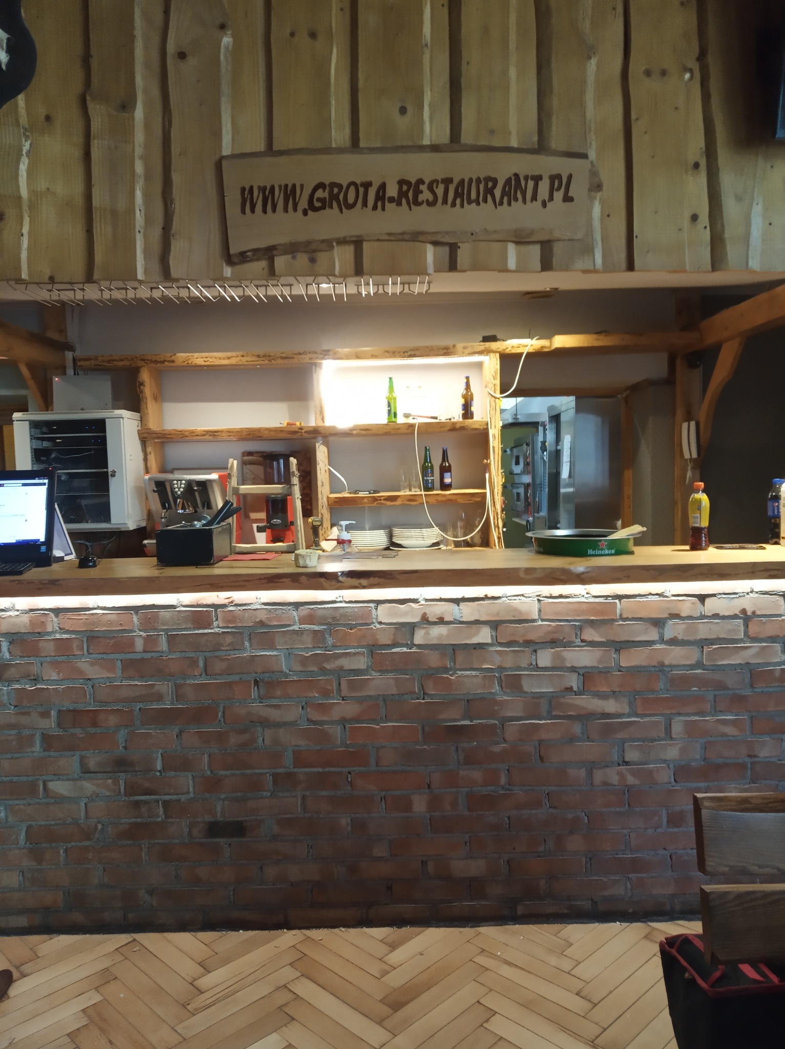 Bar w restauracji w Rzeszowie z ceglaną ścianą, drewnianym blatem, półkami z napojami i sprzętem, oraz szyldem z adresem strony internetowej.