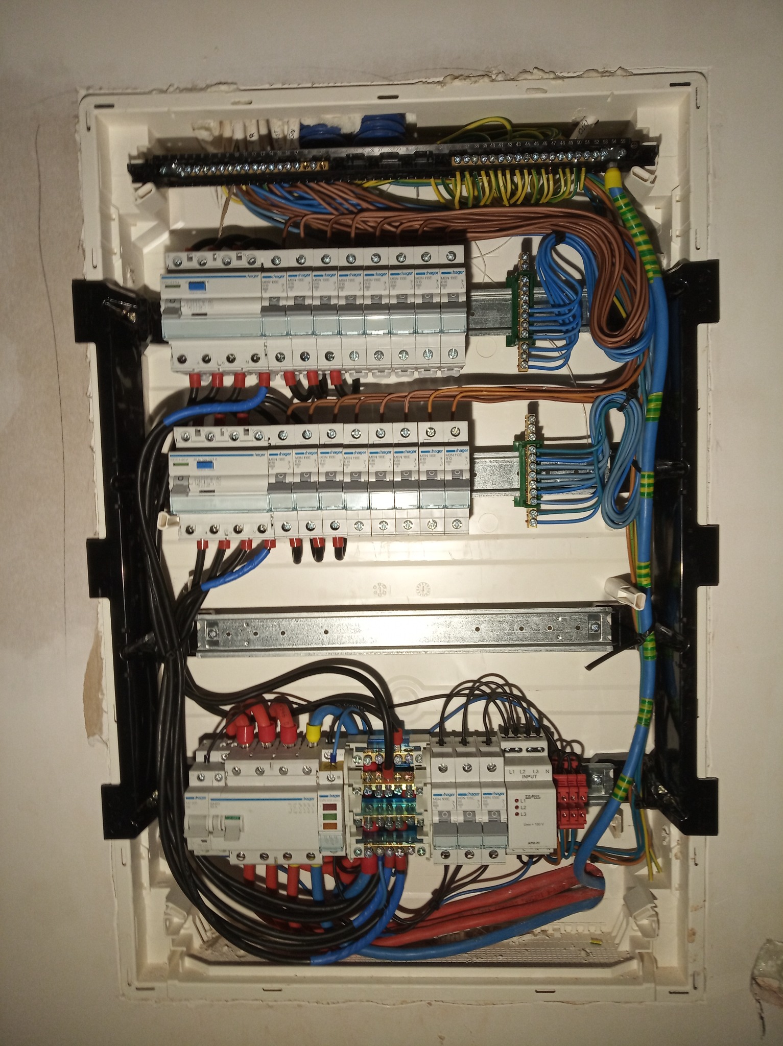 Rozdzielnia elektryczna w białej obudowie z widocznymi bezpiecznikami, przewodami o różnych kolorach (niebieski, brązowy, czarny, czerwony) i listwami zaciskowymi, zamontowana na ścianie...