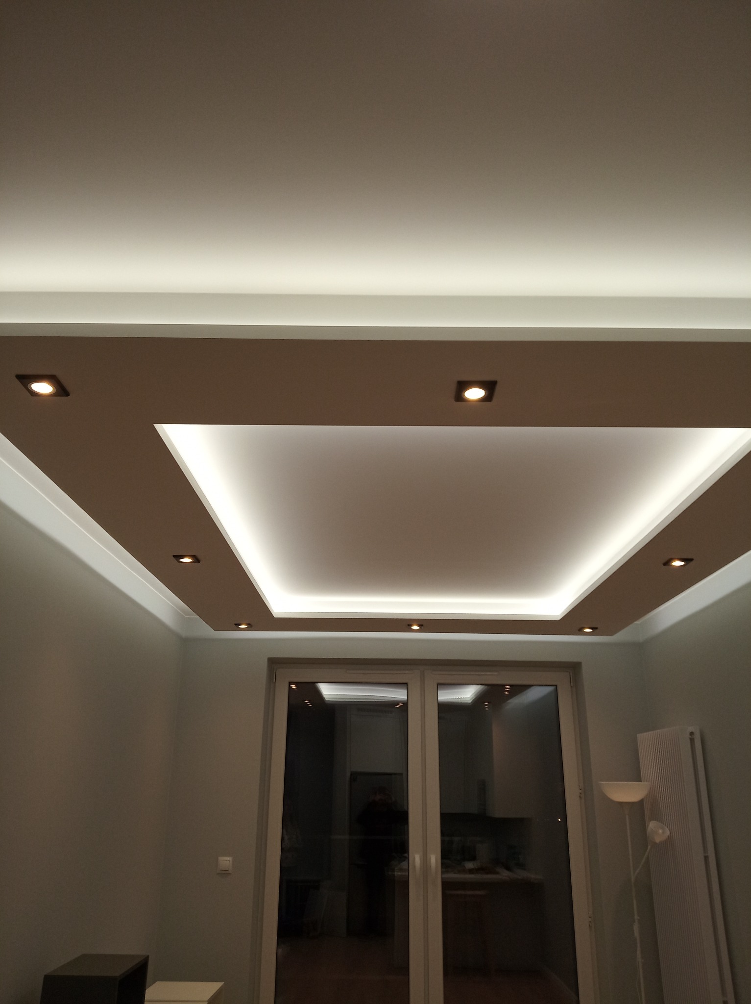 Podwieszany sufit z oświetleniem LED punktowym i taśmowym, widok z dołu na nowoczesny design w jasnym salonie z widocznym fragmentem okna balkonowego.