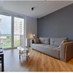 Jasne wnętrze pokoju dziennego po remoncie: szara ściana, drewniana podłoga, okno balkonowe z widokiem na budynek mieszkalny, szara sofa i szklany stolik kawowy.