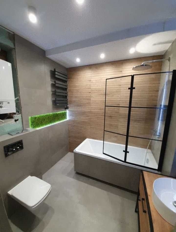 Nowoczesna łazienka z wanną z czarną szybą, umywalką na drewnianej szafce, toaletą podwieszaną i panelem z zielonego mchu.