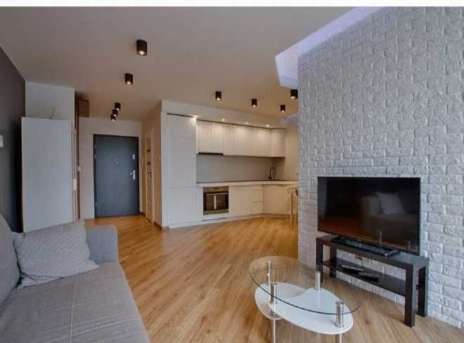 Jasne wnętrze mieszkania po remoncie: drewniana podłoga, biała ceglana ściana z telewizorem, sofa z poduszkami i szklany stolik kawowy, widok na białą kuchnię i ciemne lampy sufitowe.