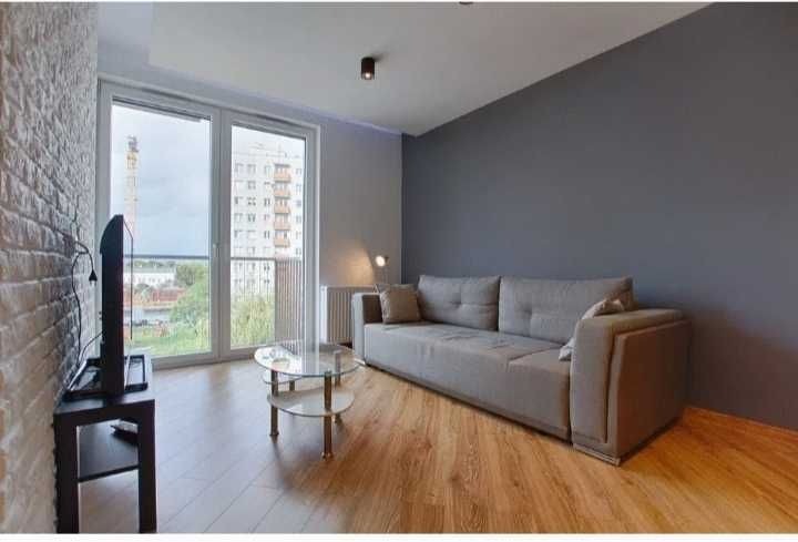 Jasne wnętrze pokoju dziennego po remoncie: szara ściana, drewniana podłoga, okno balkonowe z widokiem na budynek mieszkalny, szara sofa i szklany stolik kawowy.