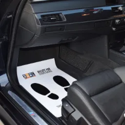 Wnętrze czarnego samochodu BMW z otwartymi drzwiami, widoczny czarny skórzany fotel, deska rozdzielcza i dywanik ochronny z nadrukiem 'Recipe for Overtaking' oraz śladami butów.