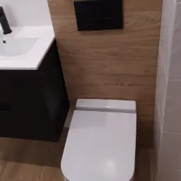 Nowoczesna toaleta podwieszana z czarnym panelem sterującym na ścianie wykończonej płytkami imitującymi drewno, obok umywalka z czarną armaturą i szafką w kolorze grafitu, całość na podłodze...