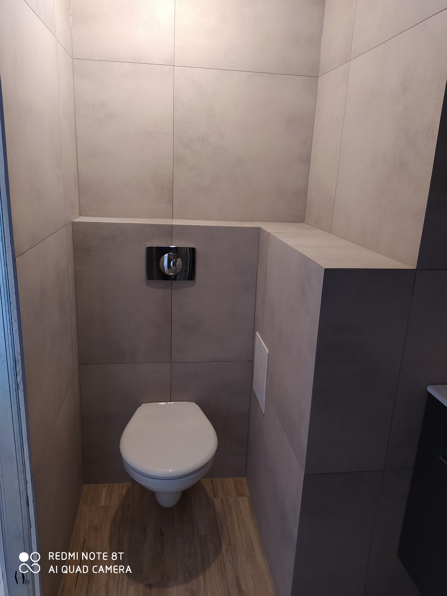 Nowoczesna, minimalistyczna toaleta w odcieniach szarości, wykończona płytkami ceramicznymi, z podwieszaną miską WC i drewnopodobną podłogą, widziana z perspektywy wejścia.