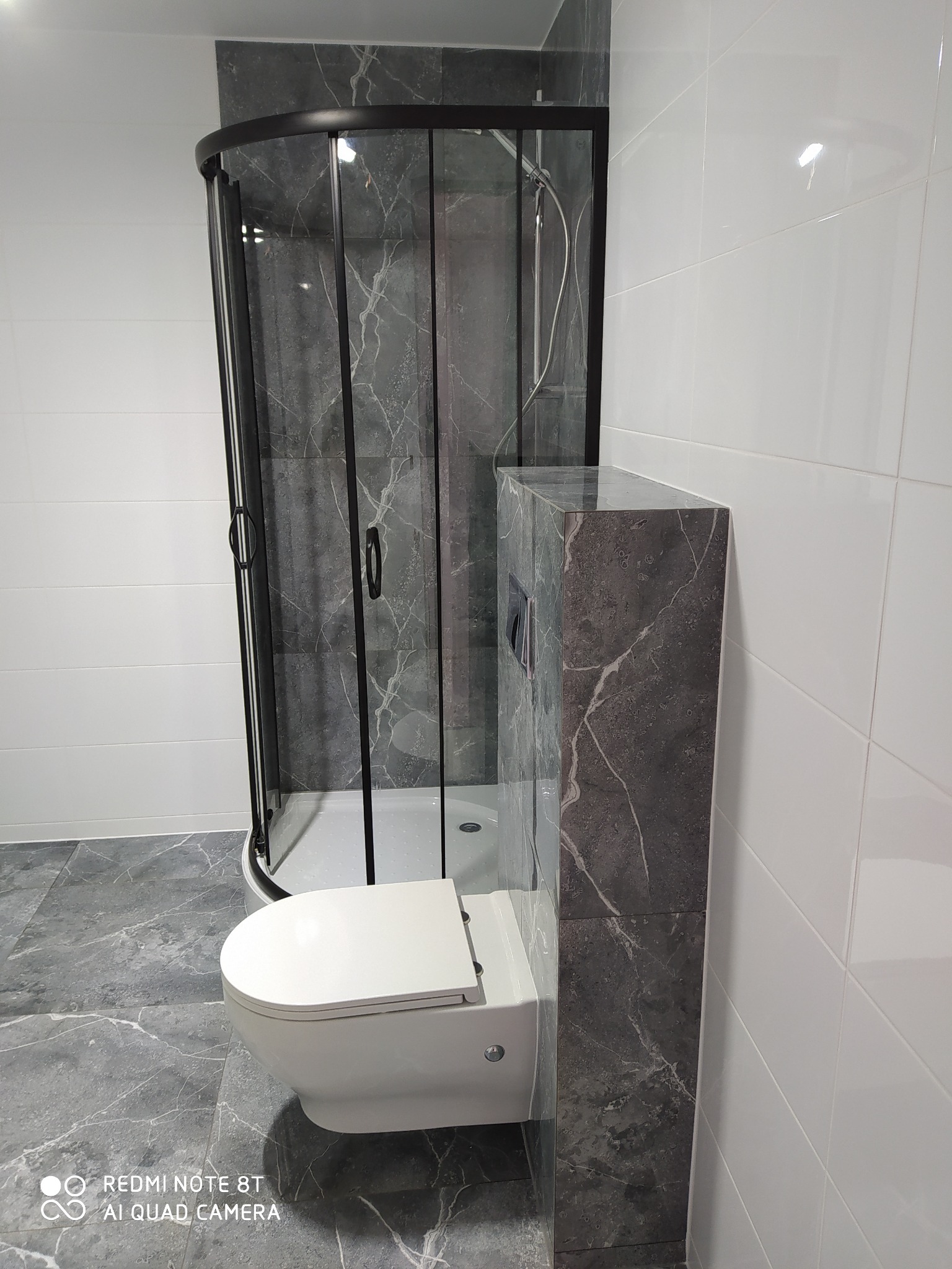 Nowoczesna łazienka z kabiną prysznicową z czarnymi ramami, podwieszaną toaletą i płytkami imitującymi marmur na podłodze i obudowie stelaża podtynkowego.