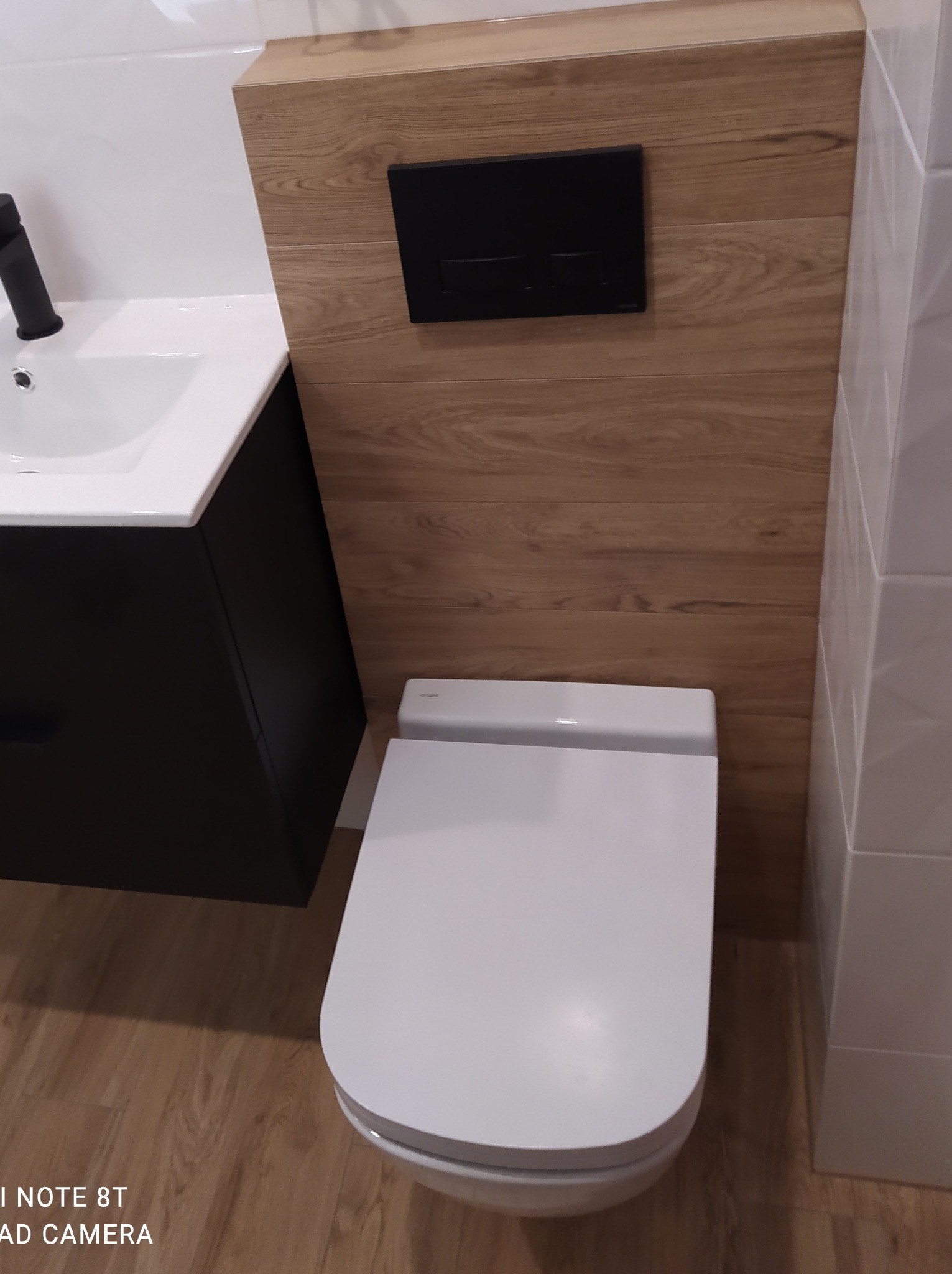 Nowoczesna toaleta podwieszana z czarnym panelem sterującym na ścianie wykończonej płytkami imitującymi drewno, obok umywalka z czarną armaturą i szafką w kolorze grafitu, całość na podłodze...