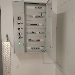 Otwarta szafa elektryczna z widocznymi bezpiecznikami, przełącznikami i trzema zielonymi diodami LED na białej ścianie. Widoczny fragment metalowej osłony i gniazdko elektryczne.