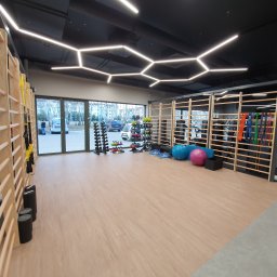 Inana - pracownia architektury wnętrz komercyjnych - Wnętrze siłowni z drewnianymi drabinkami, sprzętem do ćwiczeń, podłogą w jasnym odcieniu drewna i nowoczesnym oświetleniem sufitowym w kształcie heksagonów.