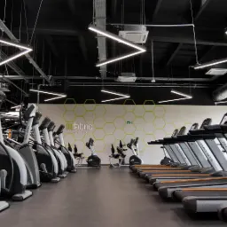 Nowoczesna siłownia FitFabric z rzędem bieżni Matrix Ultimate i orbitreków Matrix, oświetlona geometrycznymi lampami LED pod czarnym sufitem, z logo firmy na ścianie w tle.