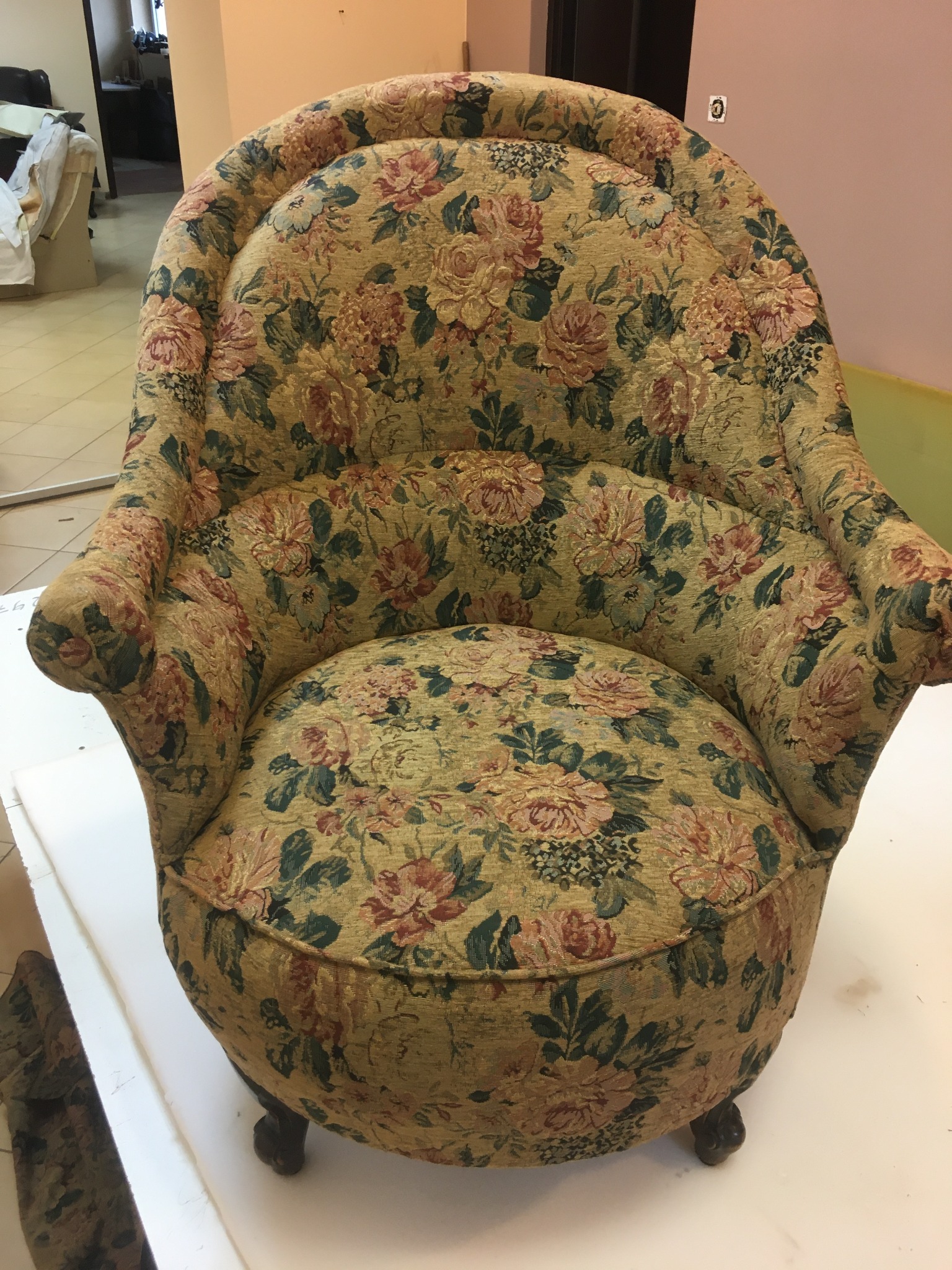 Okrągły fotel tapicerowany w kwiaty, z niskimi drewnianymi nóżkami, sfotografowany z przodu, w tle ściana w odcieniach beżu i fragment wyposażenia warsztatu.