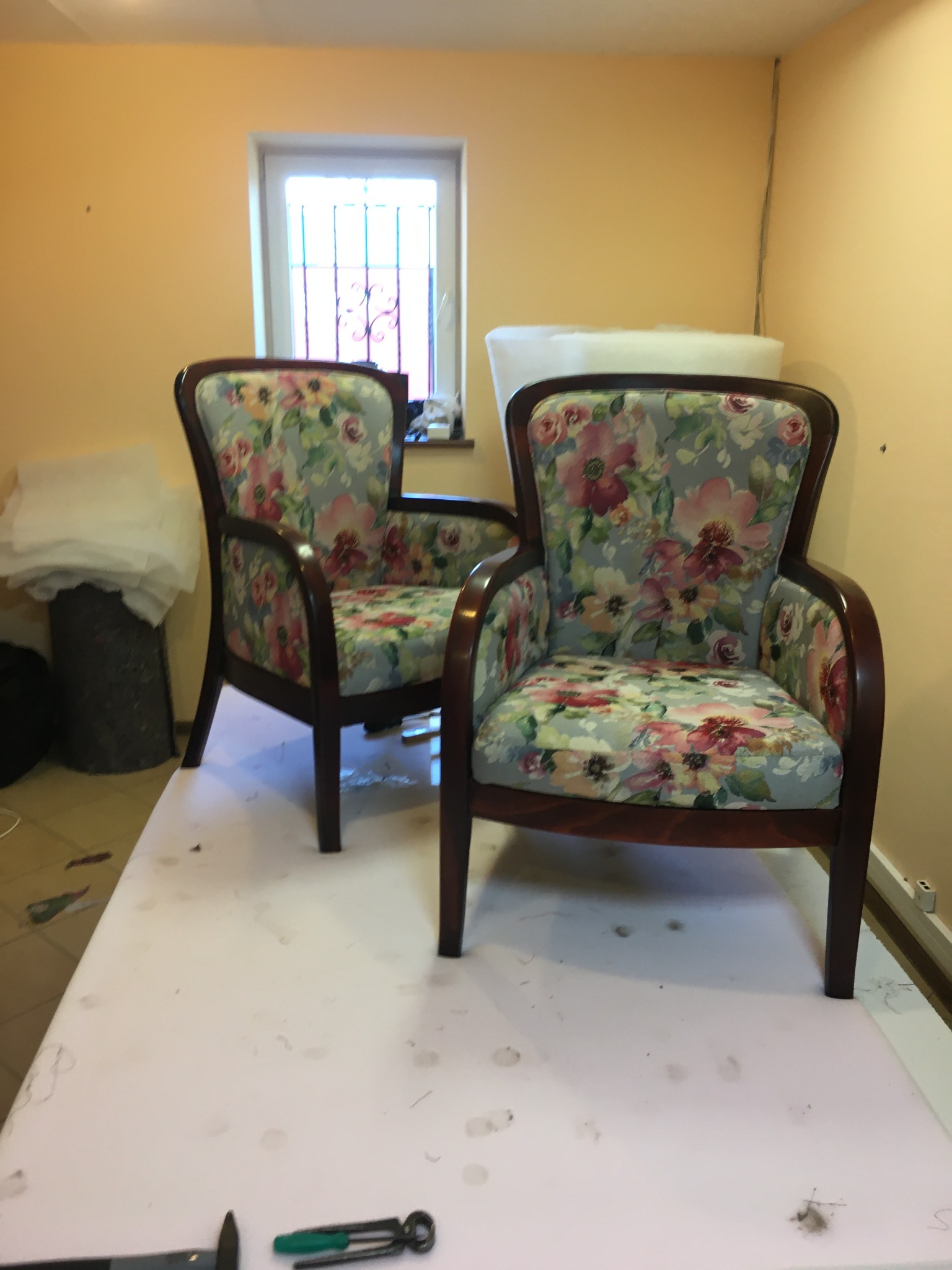 Dwa fotele po renowacji tapicerskiej z kwiatowym wzorem na szarym tle, stojące na stole roboczym w warsztacie tapicerskim, widoczne narzędzia.