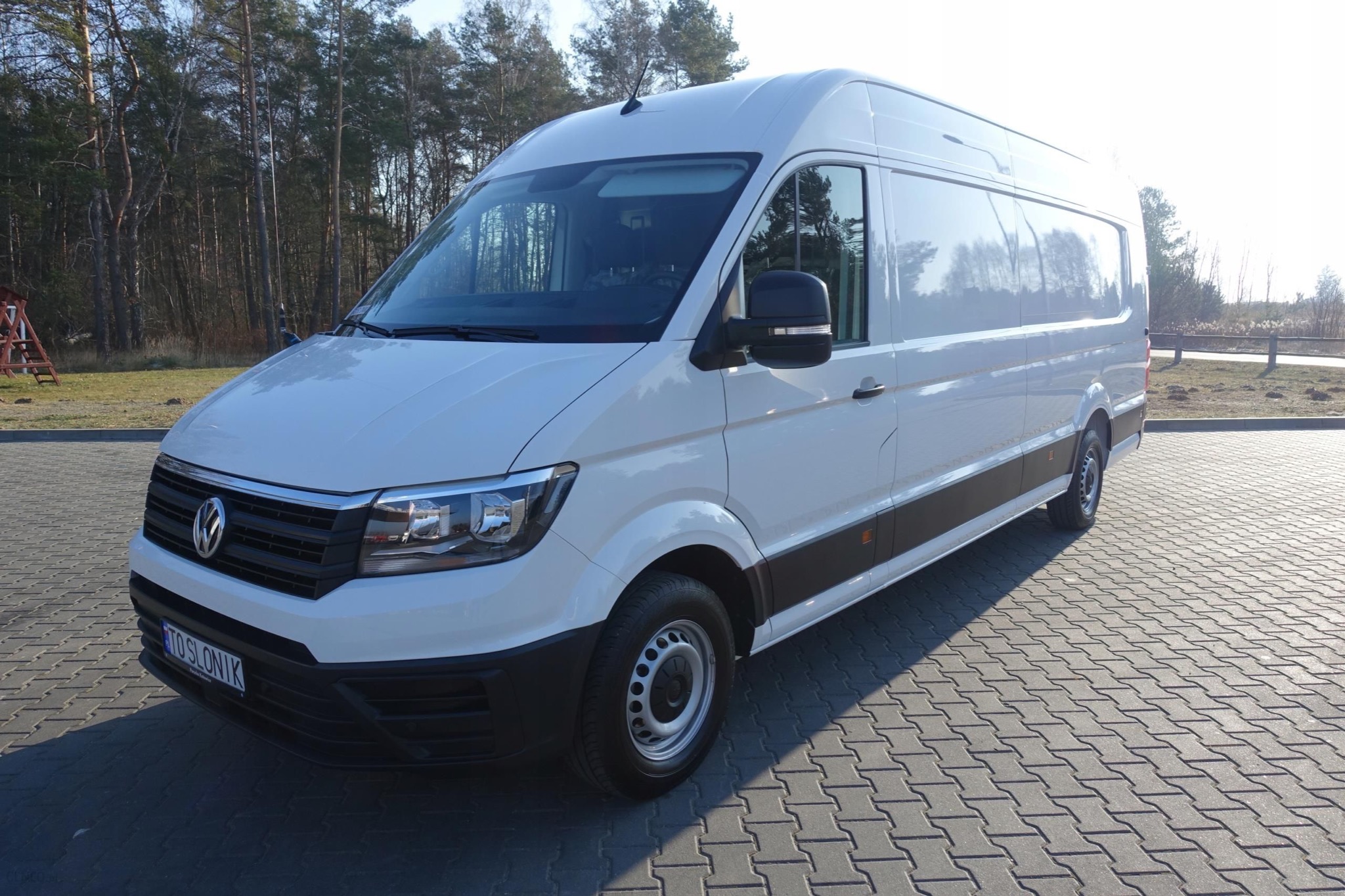 Biały bus Volkswagen Crafter z widoczną tablicą rejestracyjną 'TTO SLONIK' zaparkowany na kostce brukowej, z lasem w tle.