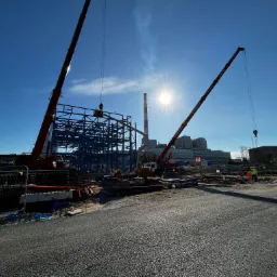 Montaż konstrukcji stalowej na placu budowy z użyciem kilku dźwigów, widoczny komin w tle i silne światło słoneczne.