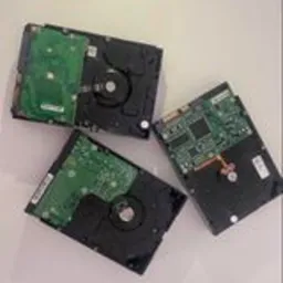 Trzy dyski twarde leżące na jasnej powierzchni, widoczne od strony elektroniki: dwa z zielonymi płytkami drukowanymi, jeden z widoczną głowicą i złączem IDE.