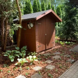 Brązowy, drewniany domek narzędziowy w ogrodzie, otoczony roślinami funkii i ścieżką z kamiennych płyt, z zegarem wiszącym na ścianie, w tle rząd strzyżonych iglaków.