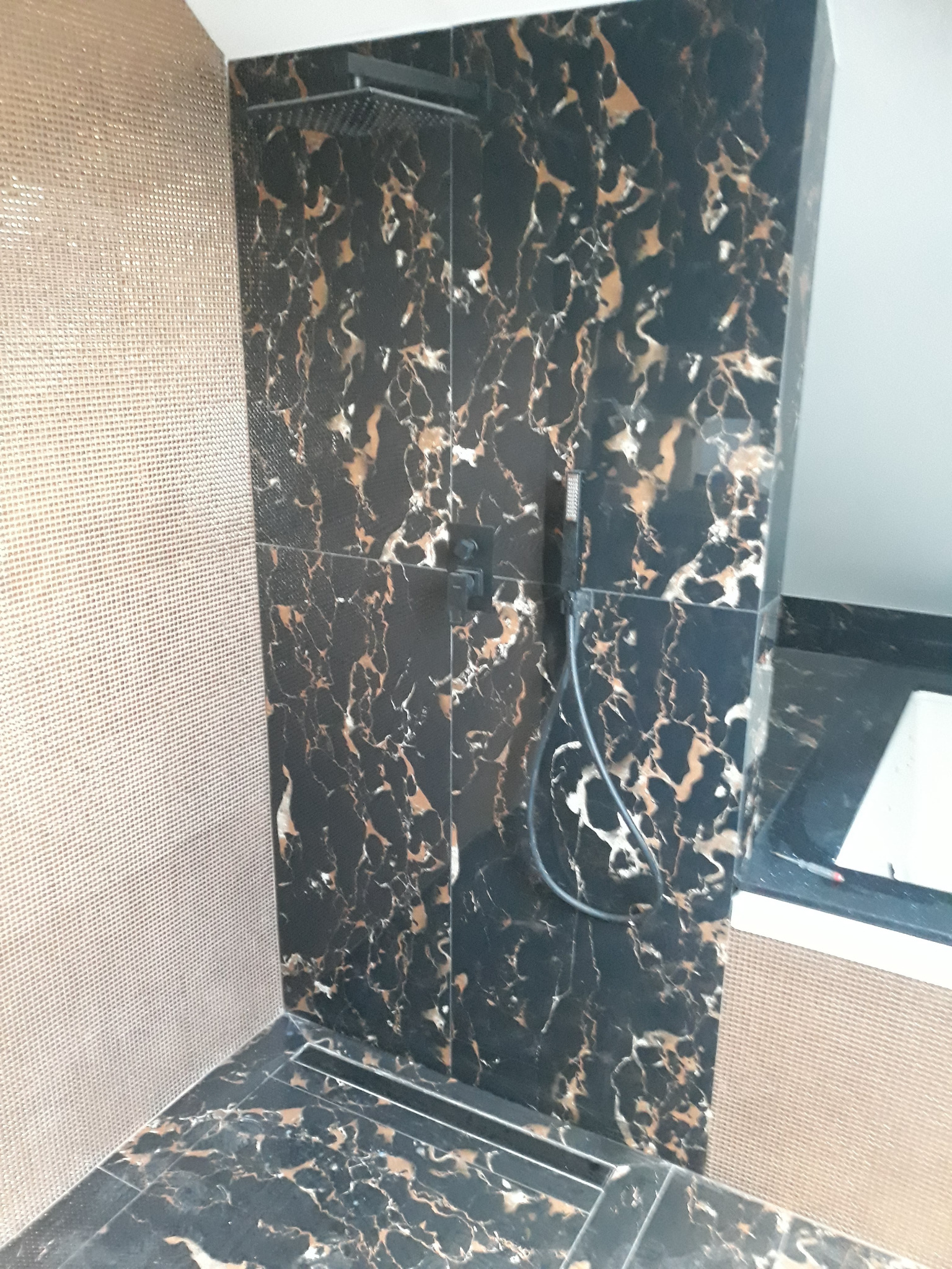 Wykończona kabina prysznicowa z czarnych płytek imitujących marmur z złotymi żyłami, sąsiadująca ze ścianą pokrytą mozaiką w odcieniach beżu, widoczny odpływ liniowy w podłodze.