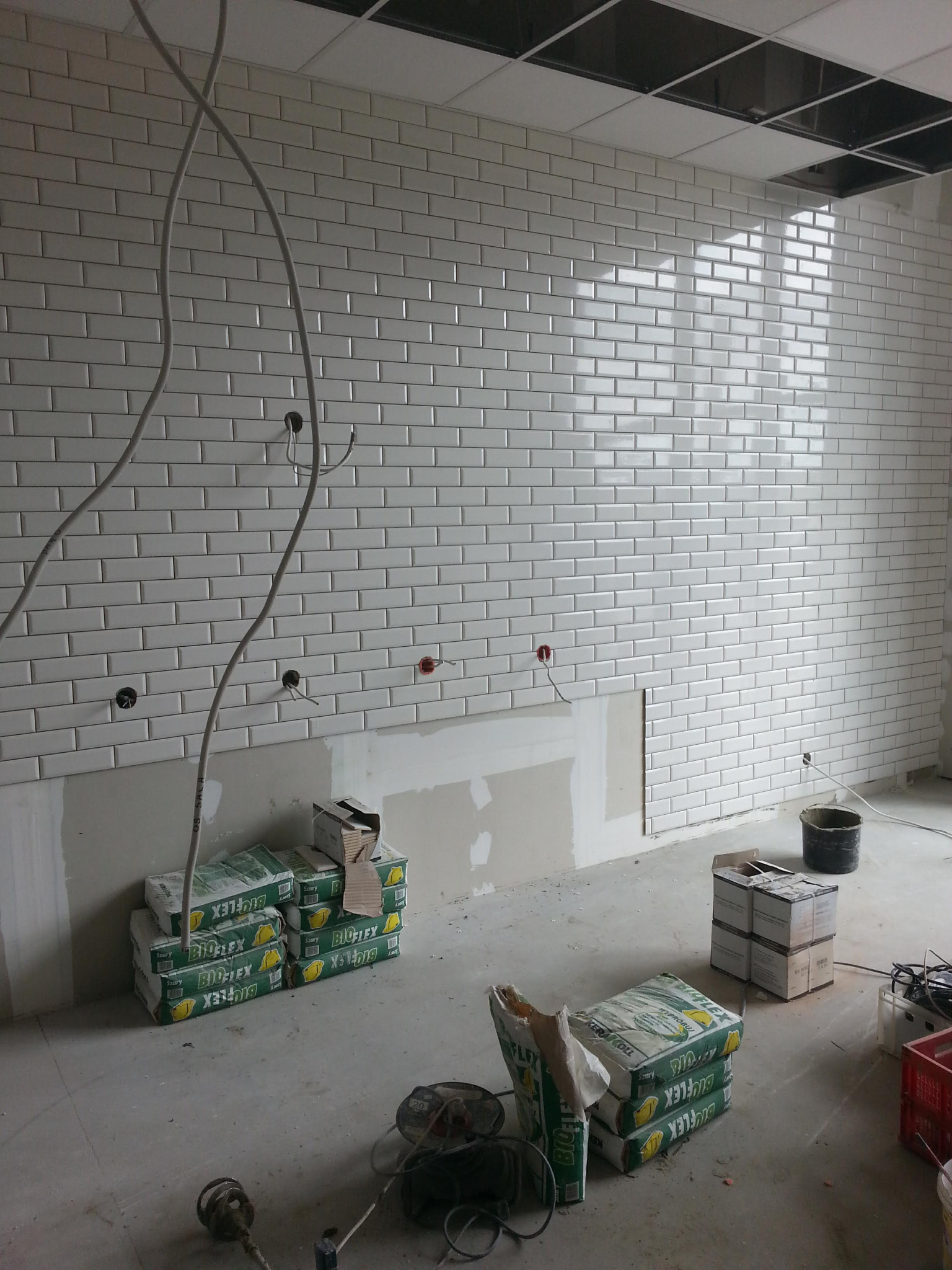 Ściana wyłożona białymi, błyszczącymi płytkami typu metro w trakcie prac wykończeniowych, widoczne puszki elektryczne, kable, worki z zaprawą klejową Bioflex i narzędzia na surowej posadzce, sufit...