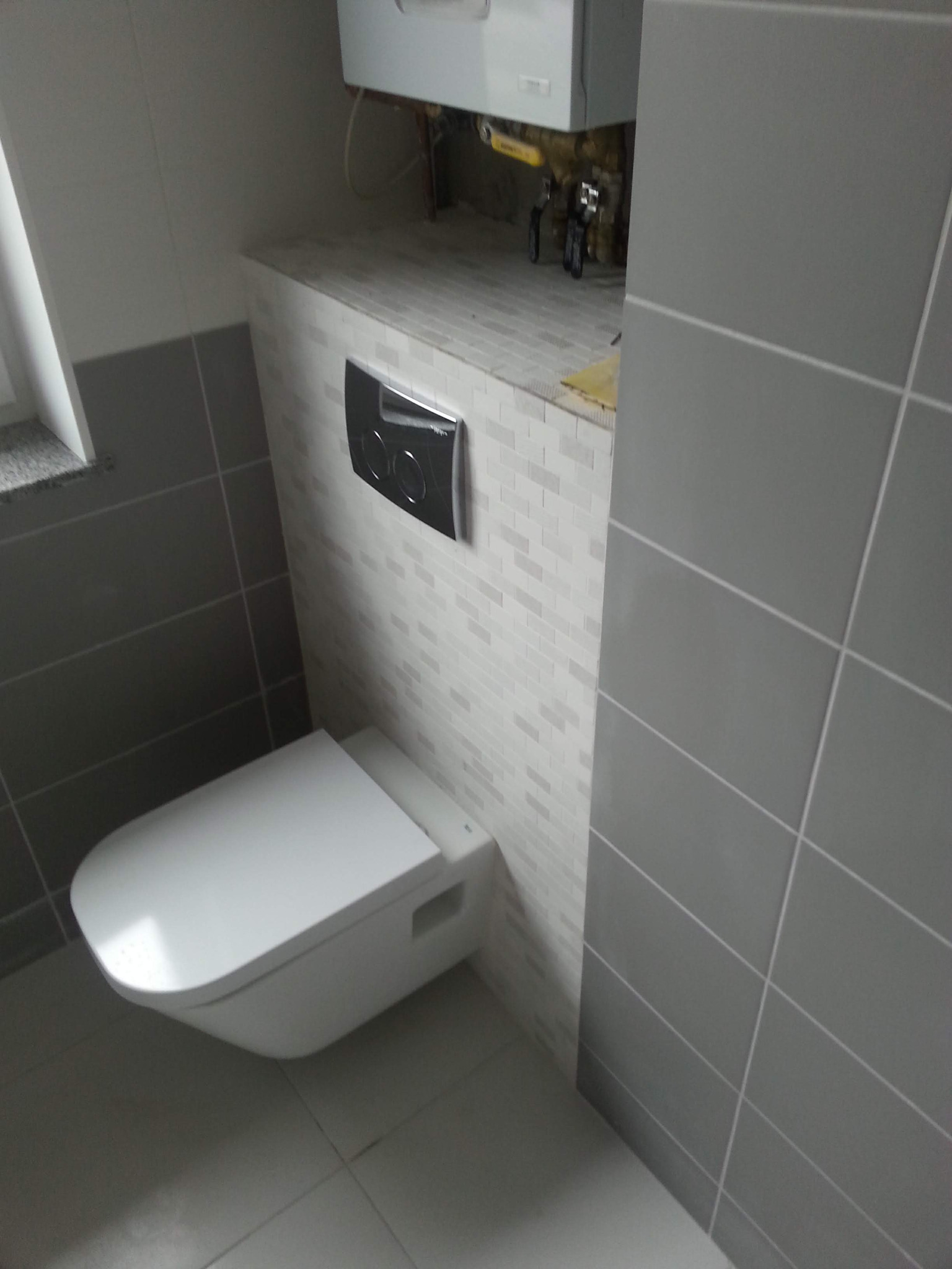 Podwieszana toaleta z chromowanym przyciskiem spłukującym na ścianie wyłożonej mozaiką, obok widoczne szare płytki. Nad toaletą ukryty pod zabudową piec gazowy.