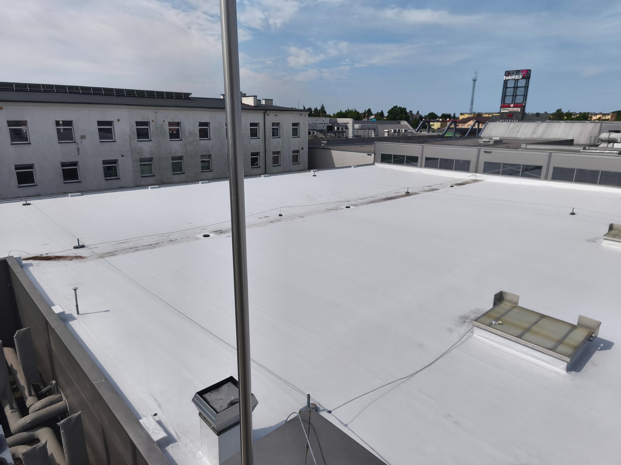 Biały, płaski dach budynku komercyjnego z widocznymi elementami wentylacyjnymi i anteną, w tle budynki miejskie i niebo z chmurami.