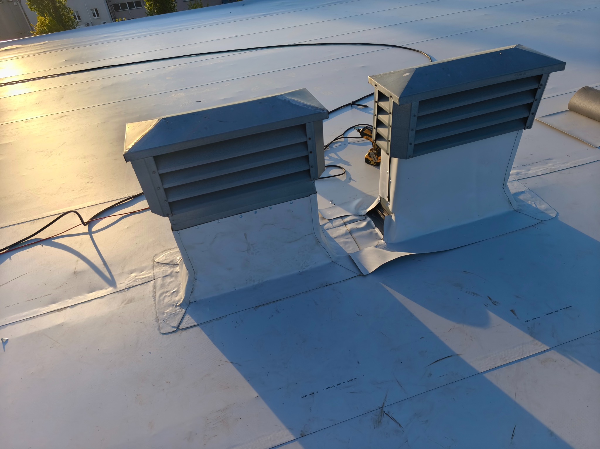 Szary dach pokryty membraną z dwoma kominami wentylacyjnymi i wkrętarką. Widoczne detale łączeń. Słoneczne światło podkreśla fakturę dachu.