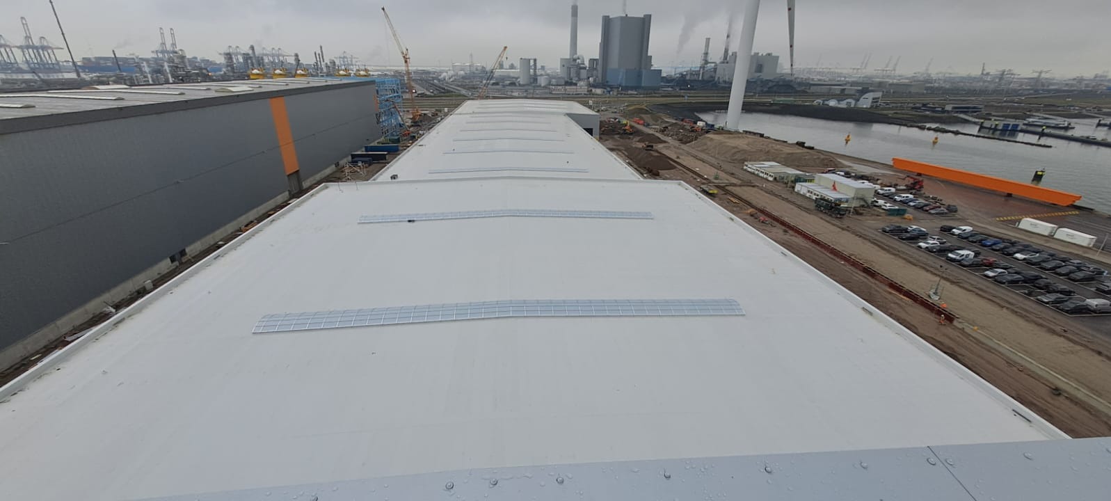 Jasnoszary dach przemysłowy z świetlikami, widziany z góry, w tle port z dźwigami i kominami. Nowoczesne pokrycie, równa powierzchnia.
