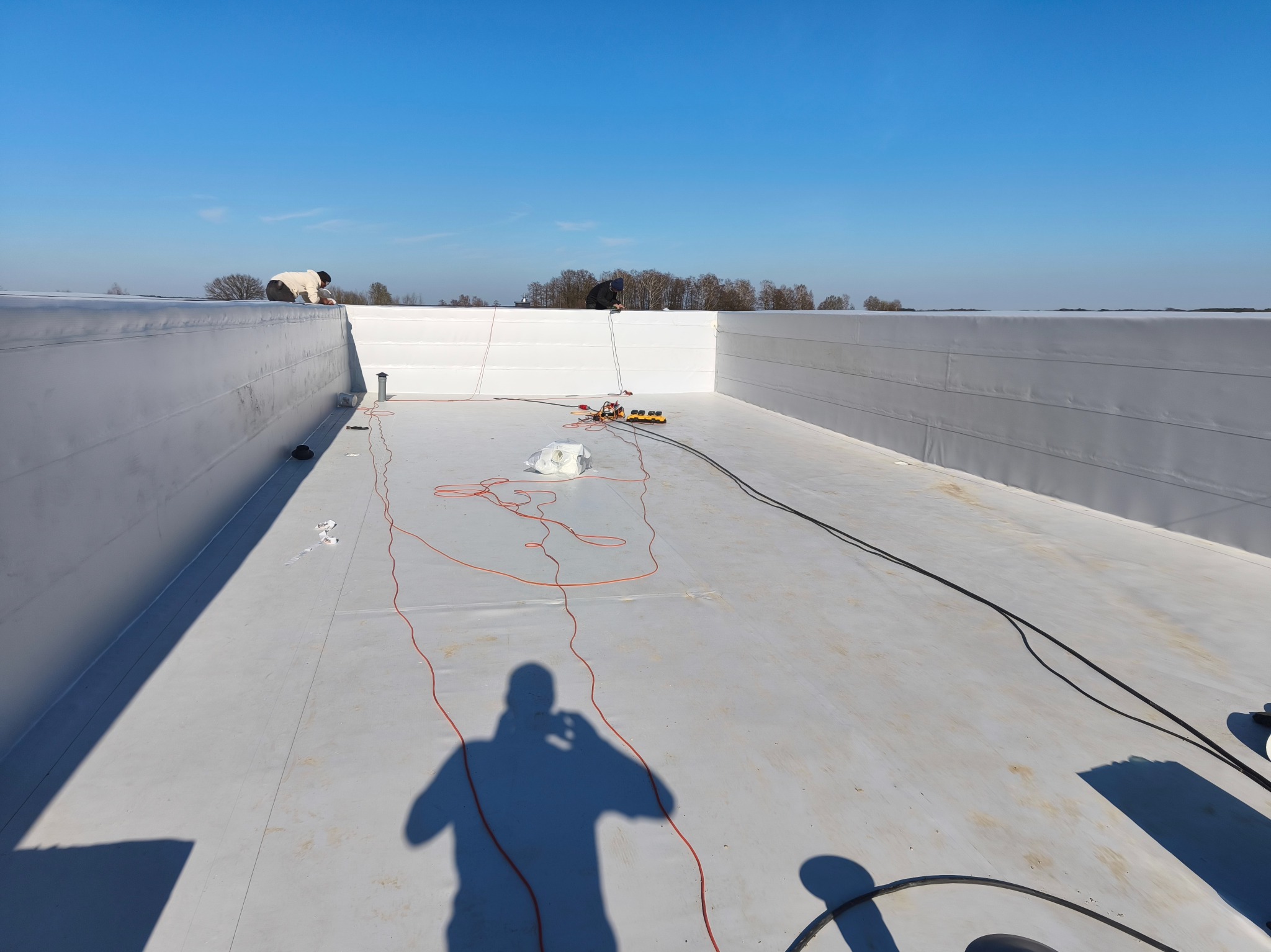 Białe poszycie dachu z widocznymi pracownikami i narzędziami, wykonane w słoneczny dzień. Cień osoby robiącej zdjęcie na pierwszym planie.