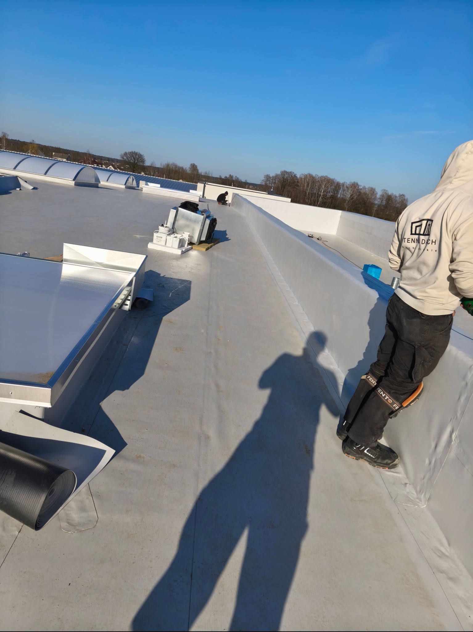 Prace na dachu płaskim: dekarz w odzieży roboczej, rozwijana rolka izolacji, widoczne elementy wentylacji. Słoneczny dzień, błękitne niebo w tle.