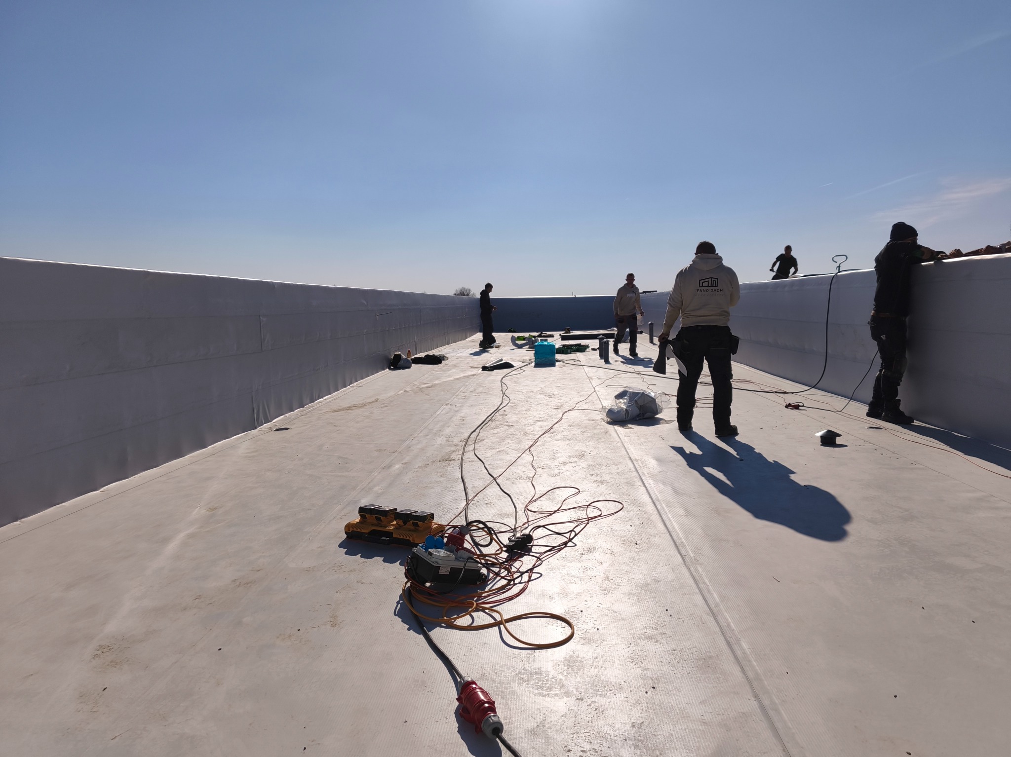 Ekipa dekarzy na płaskim dachu z membraną, narzędzia i kable na powierzchni, błękitne niebo w tle. Widok z perspektywy szerokokątnej.