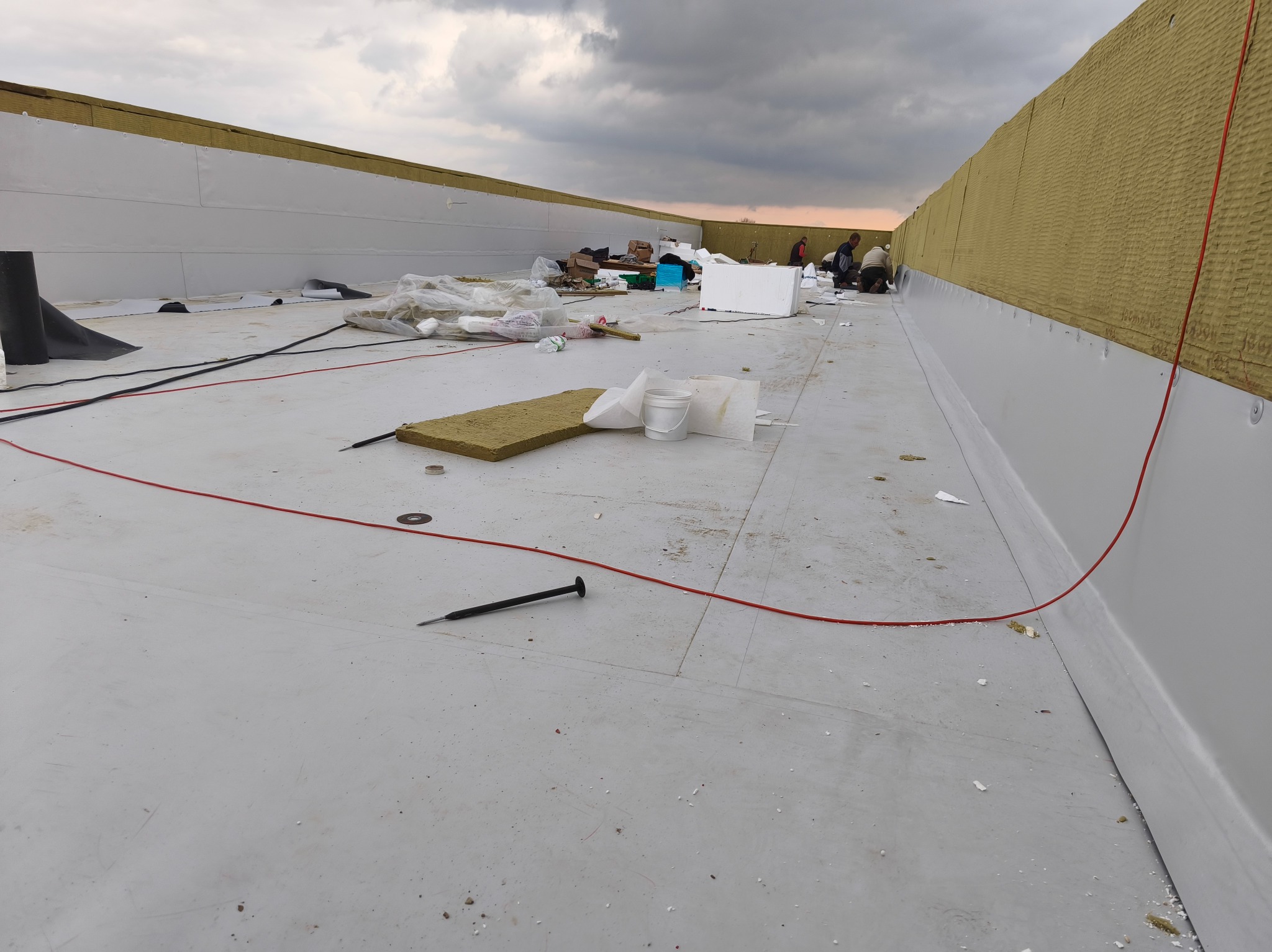 Montaż izolacji na dachu płaskim z widocznymi pracownikami i materiałami budowlanymi. Widok z góry na rozległą powierzchnię dachu.