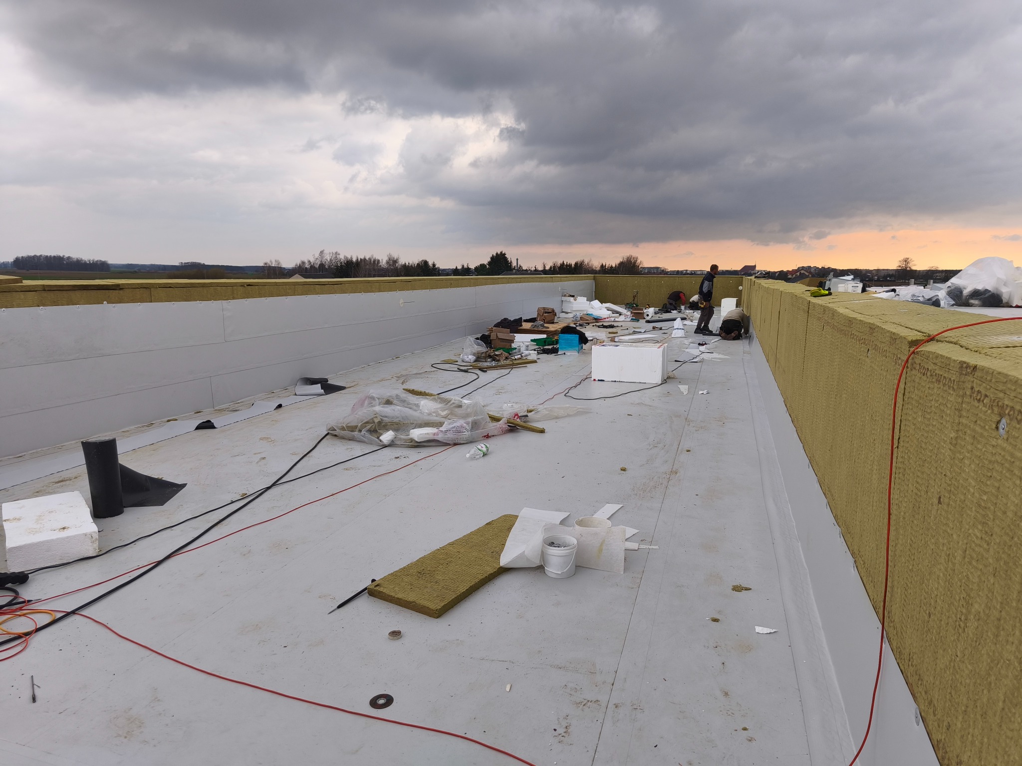 Montaż izolacji termicznej na dachu płaskim z widocznymi pracownikami, elementami instalacji i materiałami budowlanymi na tle pochmurnego nieba.