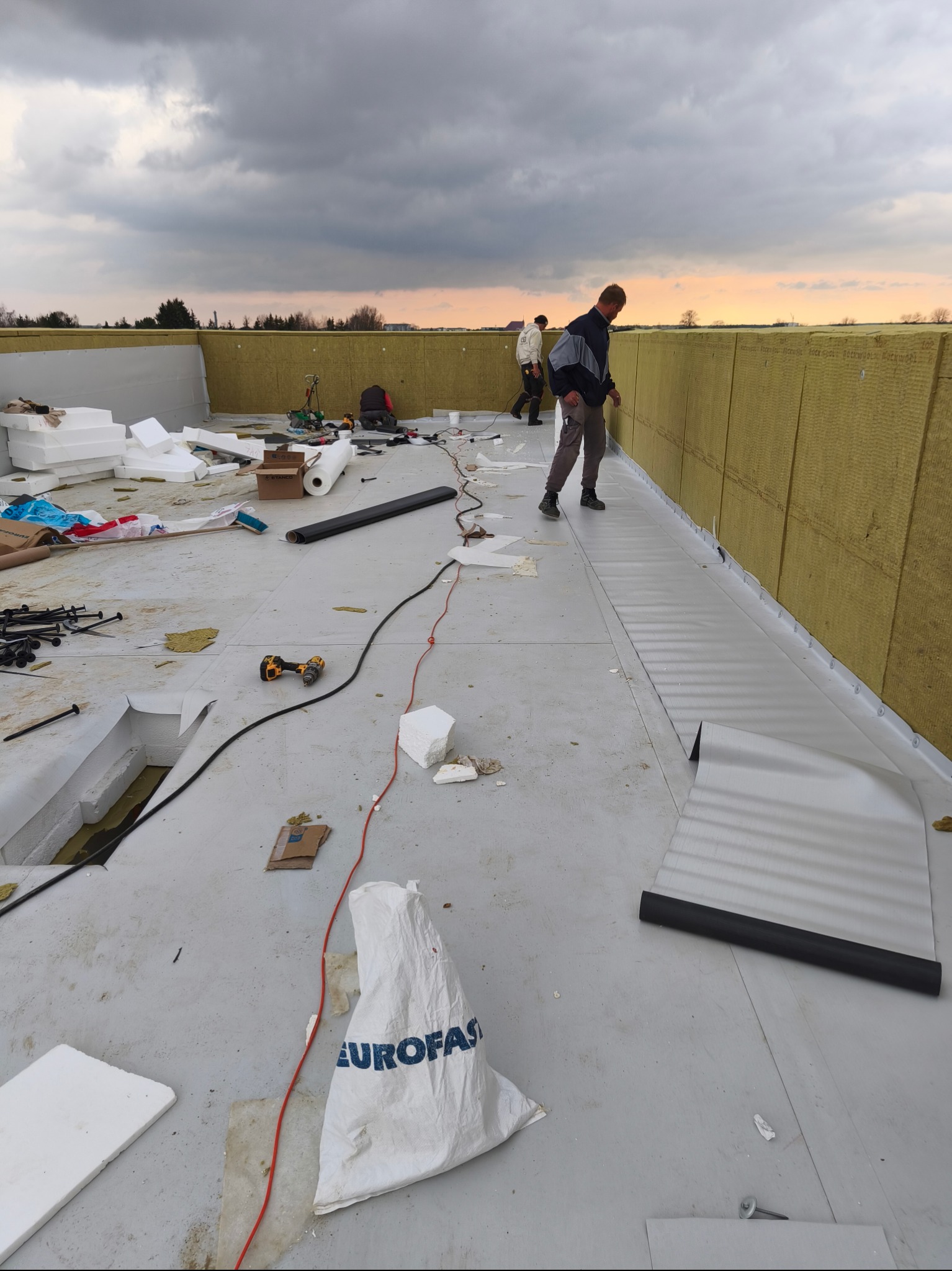 Montaż izolacji na dachu płaskim z widocznymi pracownikami, narzędziami i materiałami budowlanymi, w tle pochmurne niebo o zmierzchu.