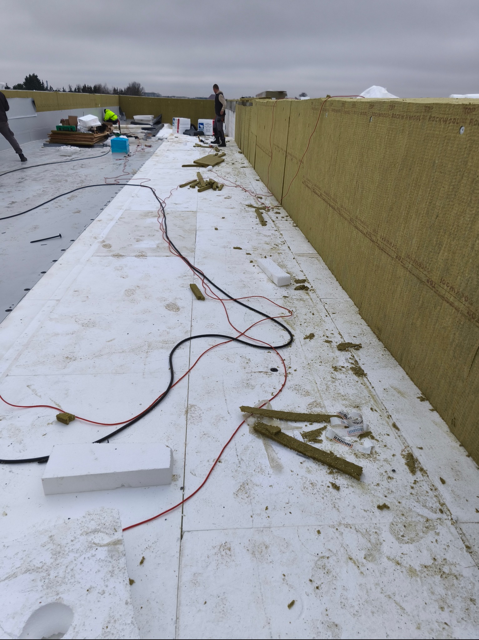 Montaż izolacji termicznej z wełny mineralnej na dachu płaskim. Widoczne przewody i płyty styropianowe. Prace dekarskie w toku, pochmurna pogoda.