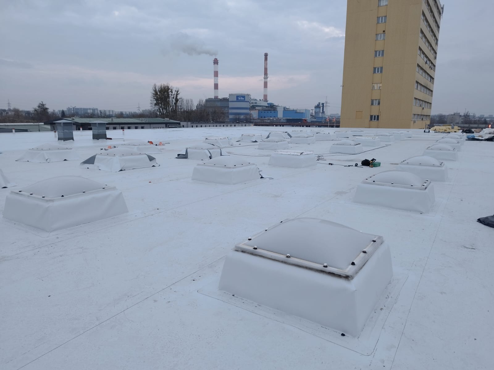 Biały dach przemysłowy z świetlikami kopułowymi, kominy w tle, budynek biurowy z prawej strony. Widok z góry na płaską powierzchnię.