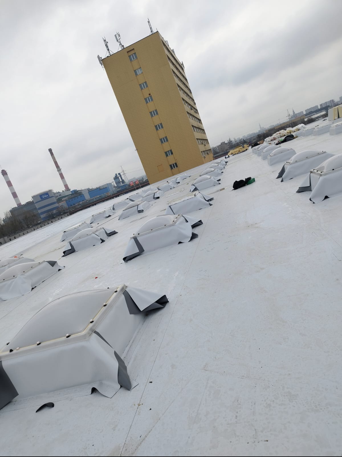 Biały dach przemysłowy z świetlikami, wysoki budynek w tle pod pochmurnym niebem. Widok z góry na płaską powierzchnię z elementami wentylacyjnymi.