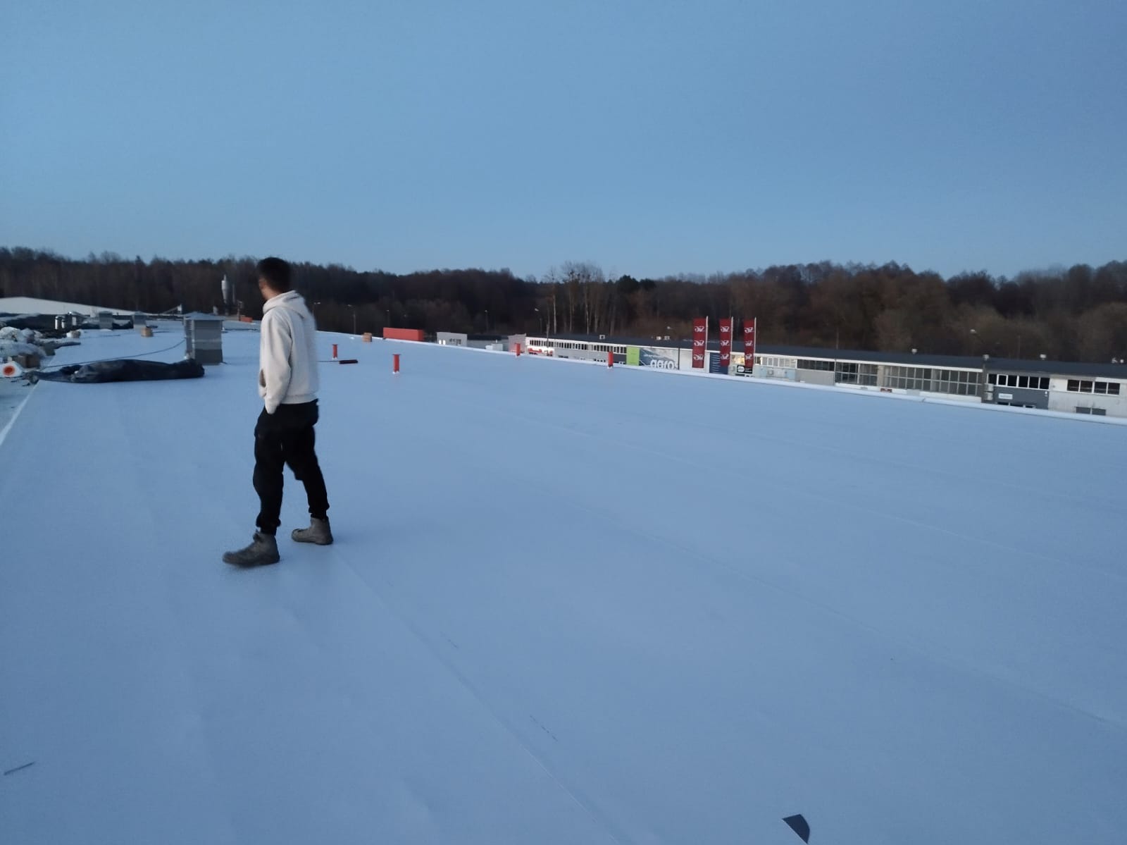 Jasny dach z białej membrany z widoczną osobą i elementami infrastruktury w tle, wykonany w pochmurny dzień.